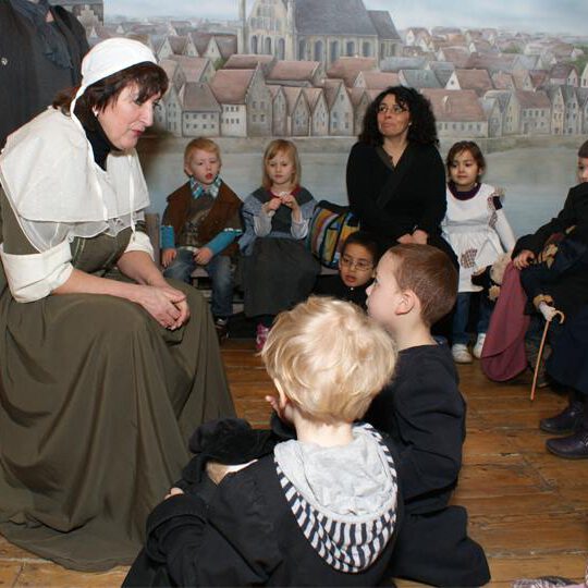 Schulklasse in der Ausstellung des Bremer Geschichtenhauses: Eine Frau in historischer Kleidung erzählt einer Gruppe von Kindern eine Geschichte. Die Kinder sitzen auf dem Holzboden, einige in historischen Kostümen. Im Hintergrund ist ein Wandgemälde mit einer Darstellung der alten Bremer Stadtansicht zu sehen.