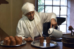 Eine Frau in historischer Kleidung, bestehend aus einem weißen Kleid und einer Haube, schenkt aus einer weißen Emaillekanne Kaffee in zwei braune Tassen ein. Die Tassen stehen auf einem silbernen Tablett. Im Hintergrund sind ein Fenster mit Blick ins Grüne und Teile eines alten Kaffeehauses zu sehen.