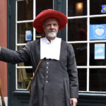 Ein Darsteller in historischer Kleidung, der eine Stadtwache darstellt, steht mit einer Hellebarde vor dem Eingang des Bremer Geschichtenhauses. Die Kulisse zeigt eine grün gerahmte Tür und Fenster. Der rote Hut und die historische Kleidung verleihen der Szene eine authentische Atmosphäre.