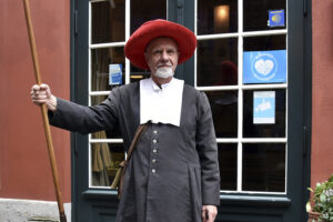 Ein Darsteller in historischer Kleidung, der eine Stadtwache darstellt, steht mit einer Hellebarde vor dem Eingang des Bremer Geschichtenhauses. Die Kulisse zeigt eine grün gerahmte Tür und Fenster. Der rote Hut und die historische Kleidung verleihen der Szene eine authentische Atmosphäre.