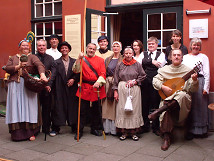 Group of performers in historical costumes in front of the entrance to the Bremen Story House. The individuals represent various historical characters, including musicians, guards, and other figures depicted at the Bremen Story House. The building's facade with windows and a door is visible in the background.