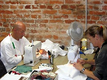 Two individuals working at sewing machines in a costume workshop. In the background, a rustic brick wall is visible. On the table are fabrics, sewing tools, and accessories while the individuals sew white fabrics.