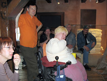 Group of individuals in a room at the Bremen Story House, including Ulrich Mickan, the original leader and organizer, standing in an orange shirt. In the foreground, a person in a wheelchair is seated, while other participants listen attentively. The room has a historical atmosphere with visible wooden beams and exhibits in the background.