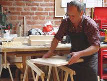 A man working in a workshop for set construction. He is wearing an apron and working on a wooden board placed on a workbench. In the background, a brick wall, tools, wood shavings, and additional materials create a creative work environment.