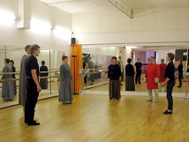 Performers rehearsing their roles in a practice room with wooden flooring and large mirrored walls. Some are dressed in historical costumes, focusing on their scenes as a group. The room is brightly lit and conveys a professional working atmosphere.