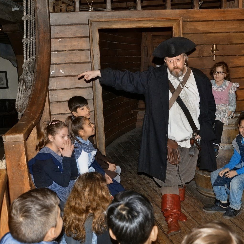 An actor dressed as a pirate gesturing and telling stories to a group of children on a wooden ship deck